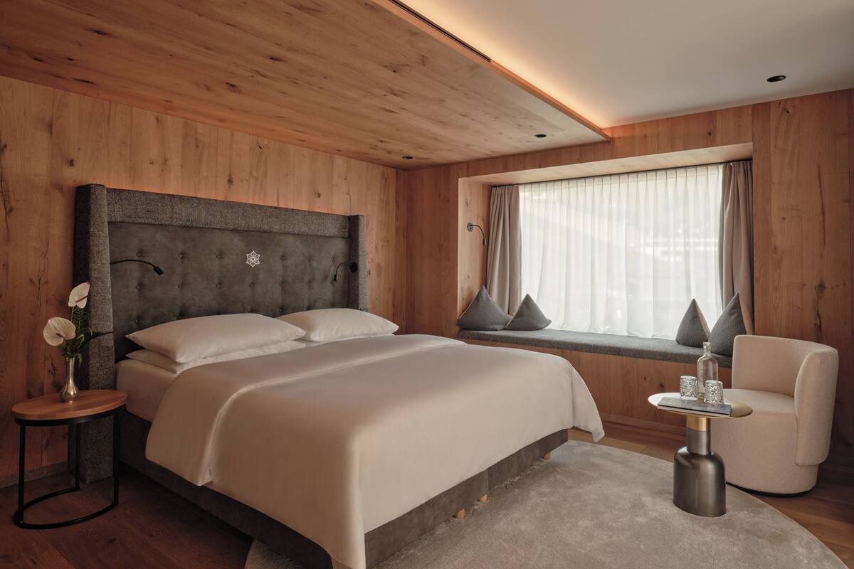 Ein modernes Schlafzimmer mit Holzbalkendecke, einem großen Bett mit weißer Bettwäsche, einem Nachttisch mit Blumen in einer Vase, einem Fenster mit Vorhängen und einem Sessel mit kleinem Tisch.