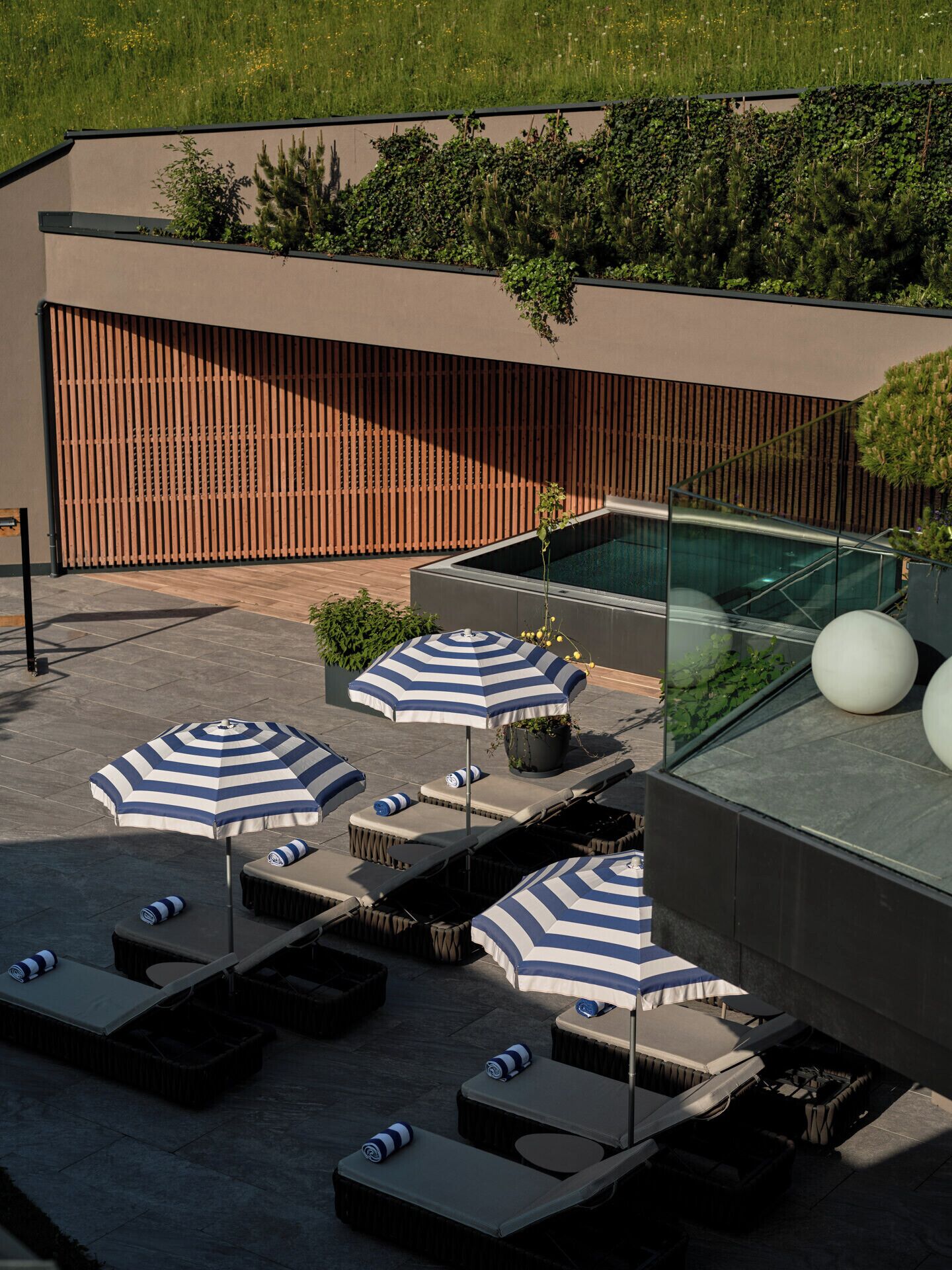 Eine Dachterrasse mit Liegestühlen, gestreiften Sonnenschirmen und einem Pool mit Glasveranda.