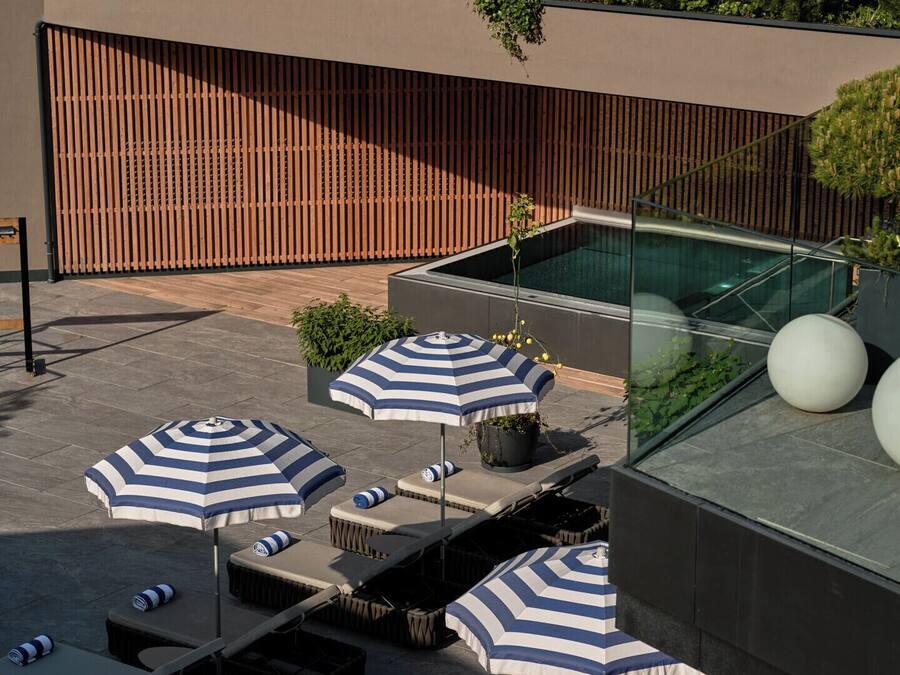Eine Dachterrasse mit Liegestühlen, gestreiften Sonnenschirmen und einem Pool mit Glasveranda.