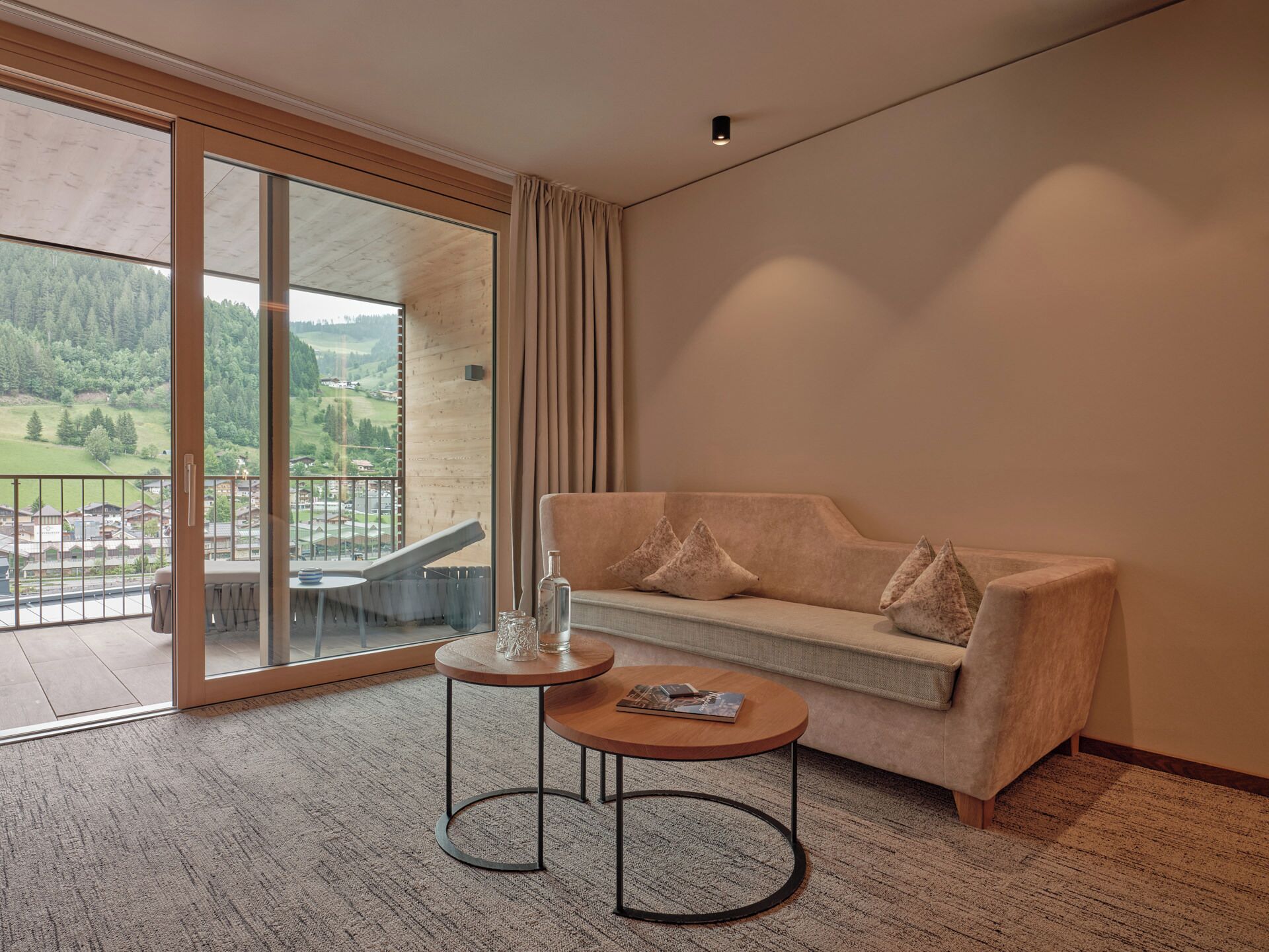 A modern hotel room with a sofa, a small round table, and a view of a green landscape through large glass doors.