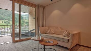 A modern hotel room with a sofa, a small round table, and a view of a green landscape through large glass doors.