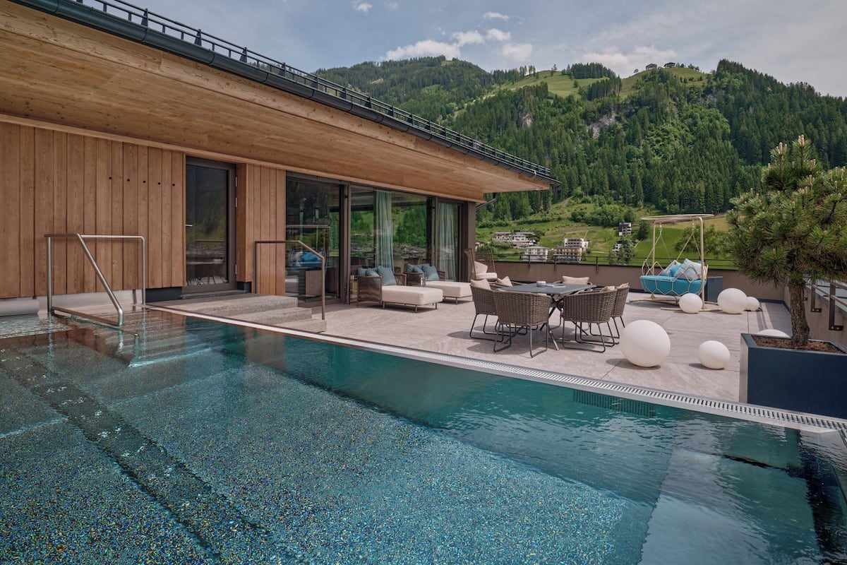 Ein moderner Poolbereich im Freien mit Holzterrasse, Sitzgelegenheiten und Blick auf eine gebirgige Landschaft.