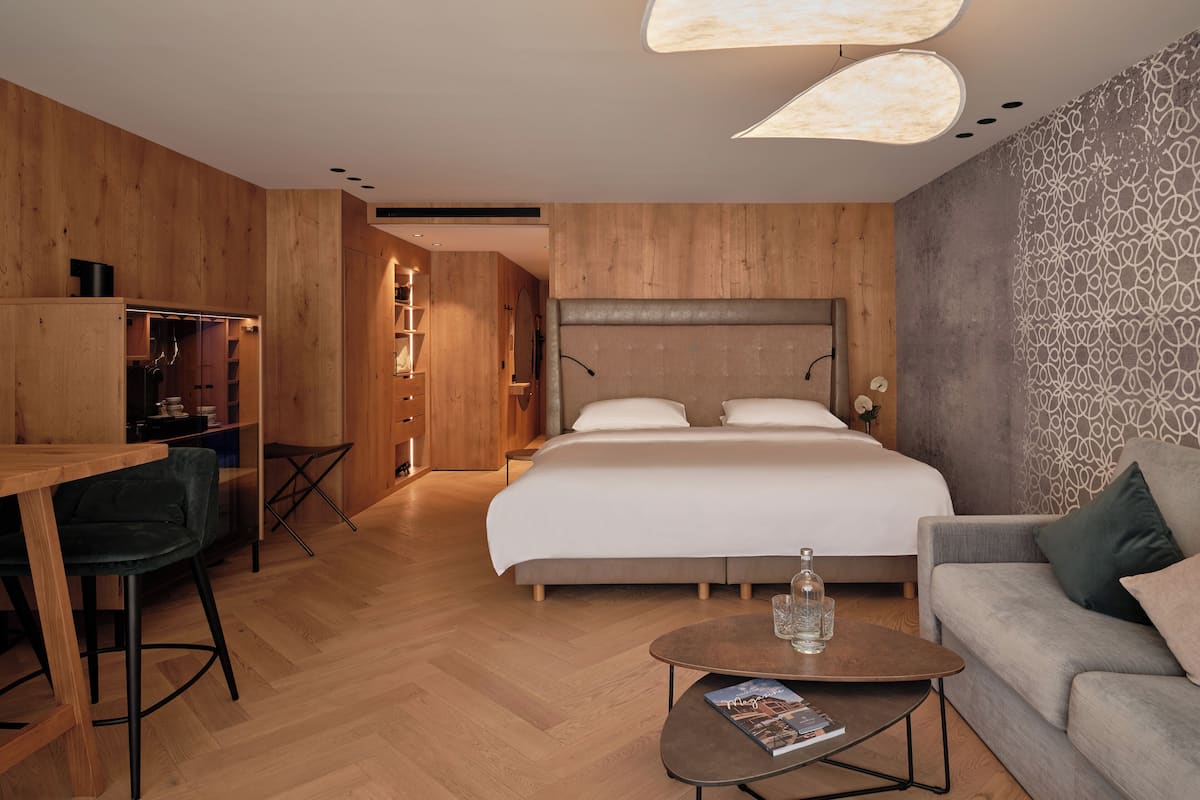 Ein modernes Hotelzimmer mit Holzboden, einem Bett mit weißer Bettwäsche, einer Couch, einem Schreibtisch mit Stuhl und einem kleinen Tisch mit einer Zeitschrift.