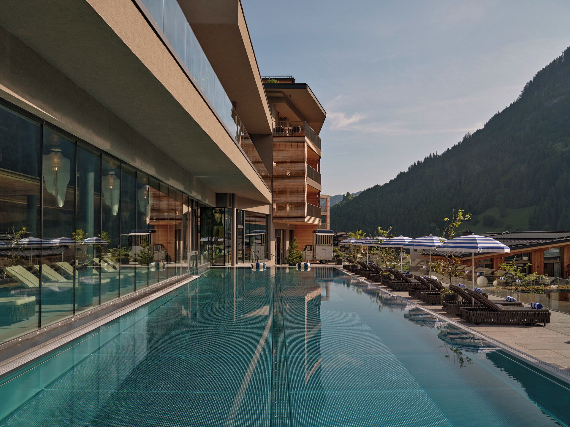 A modern hotel with a large indoor swimming pool, lounge areas with sunbeds, and a mountain backdrop.