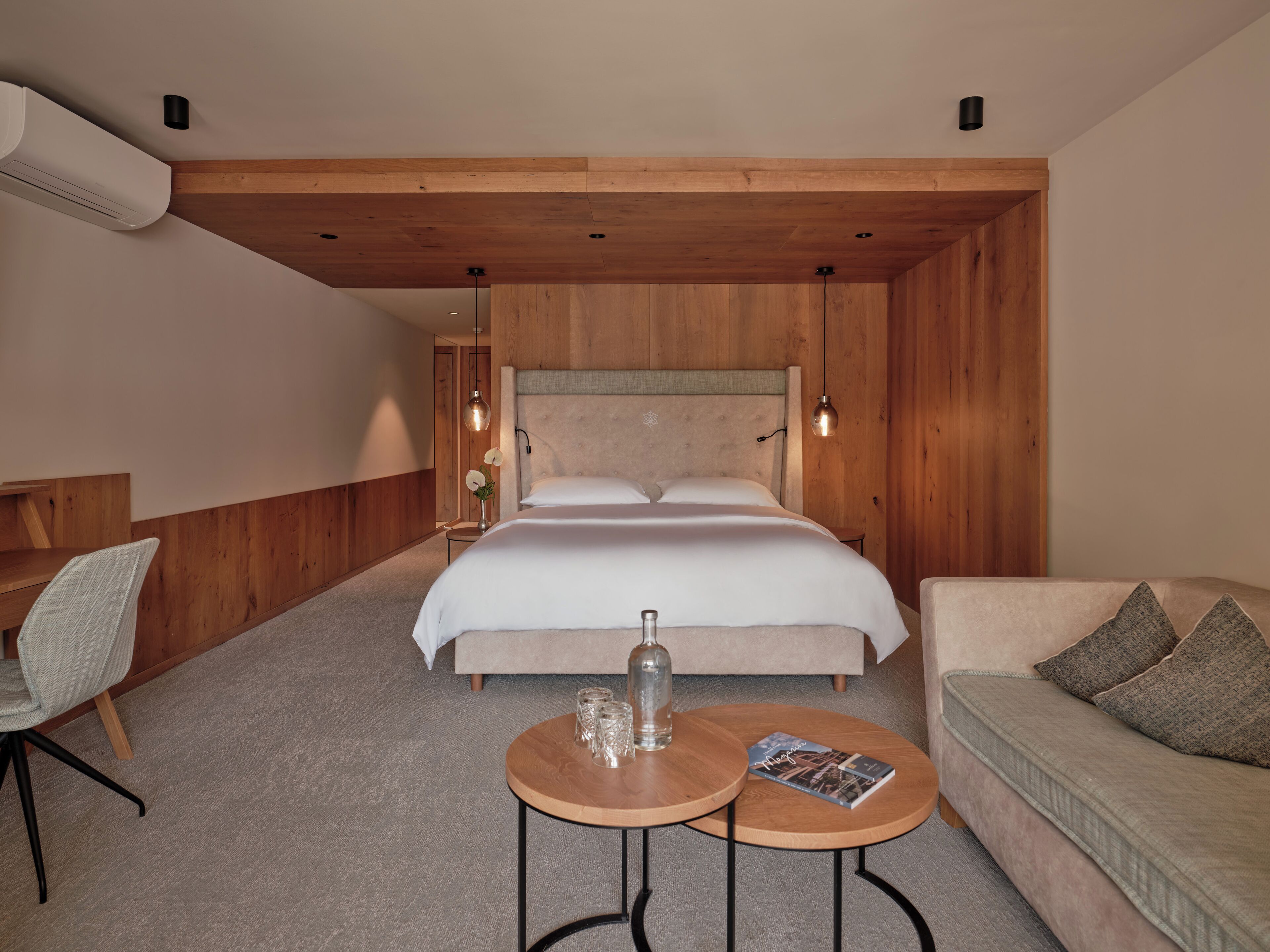 Ein modernes Hotelzimmer mit Holzboden, einem großen Bett mit weißen Leinen, einem Nachttisch aus Holz, einem runden Holztisch mit einer Zeitschrift, einer Couch mit Kissen und einem Sessel.