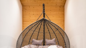 Un fauteuil suspendu doté d’un siège et d’un dossier rembourrés, fixé à une structure métallique, dans une pièce aux murs en bois et au sol carrelé.