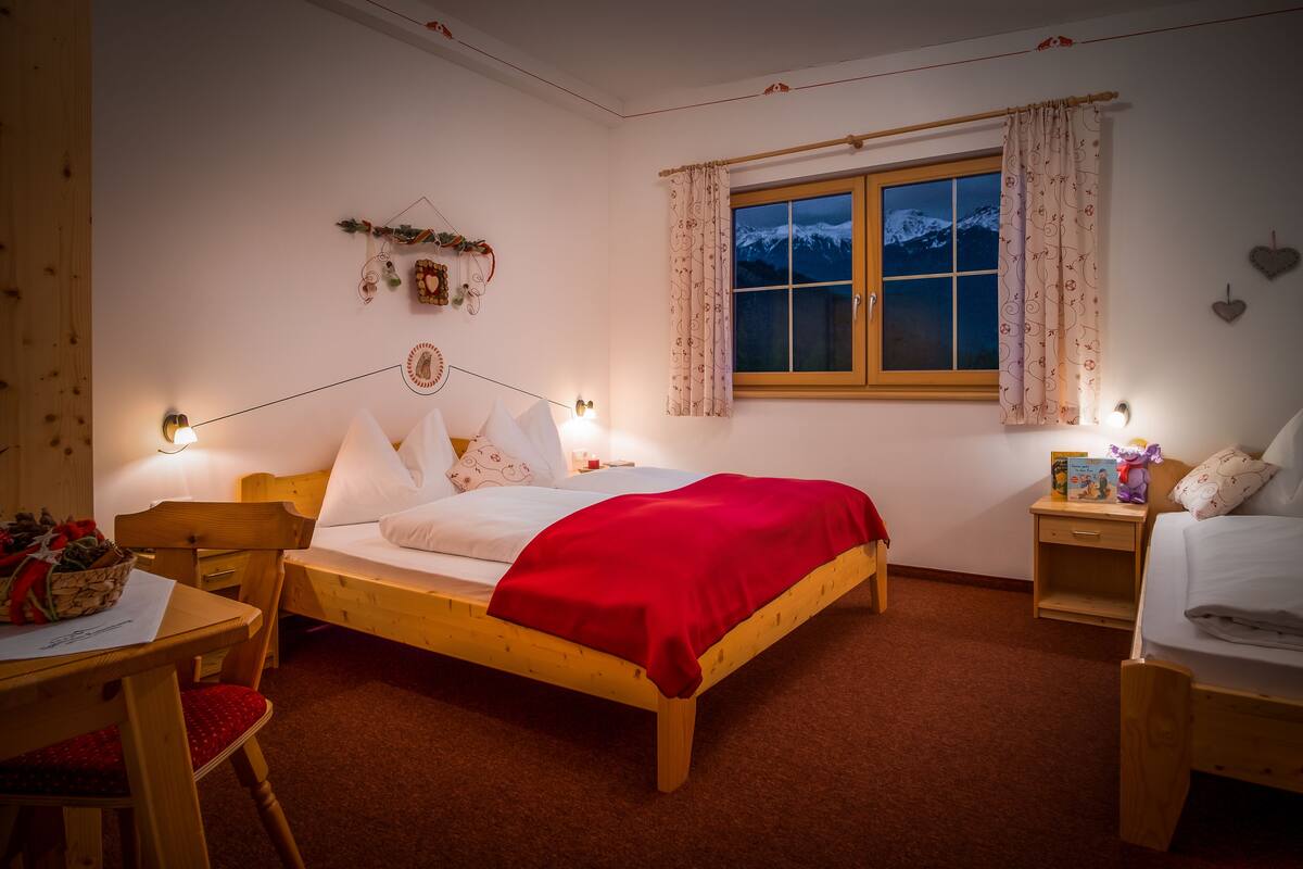 Un dormitorio acogedor con una cama de madera, un escritorio con una cesta de fruta, una mesita de noche con una lámpara y una ventana con cortinas.