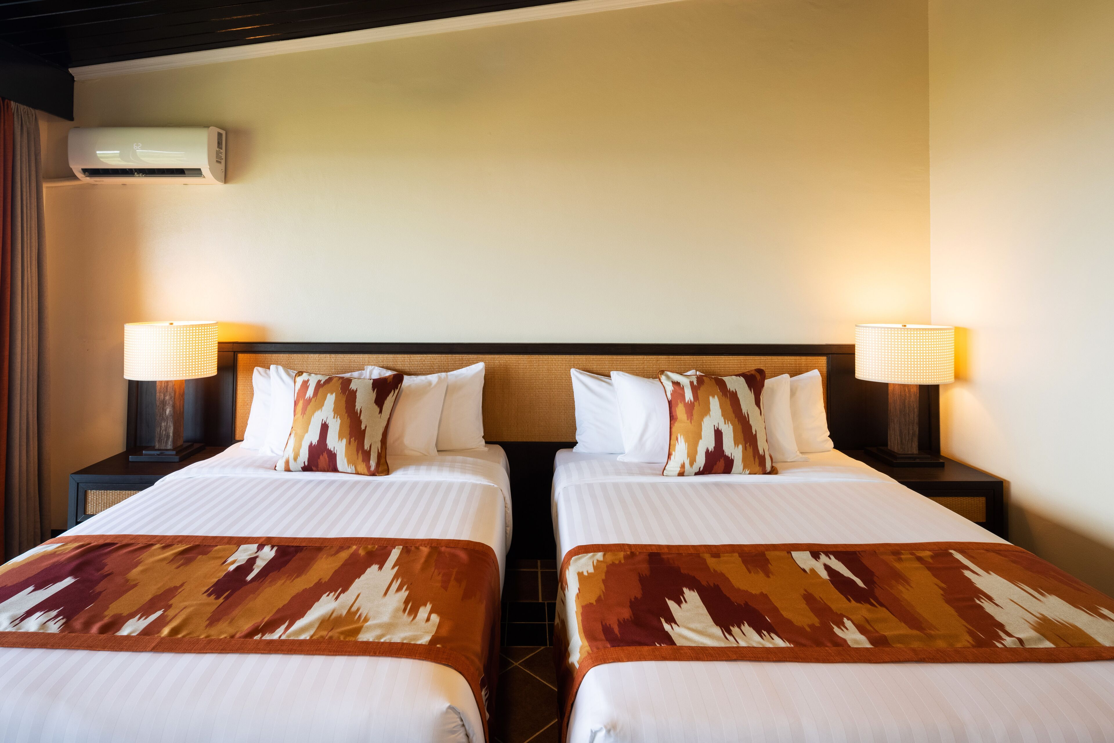 A hotel room with two beds, each with a patterned bedspread, two bedside lamps, and an air conditioning unit on the wall.