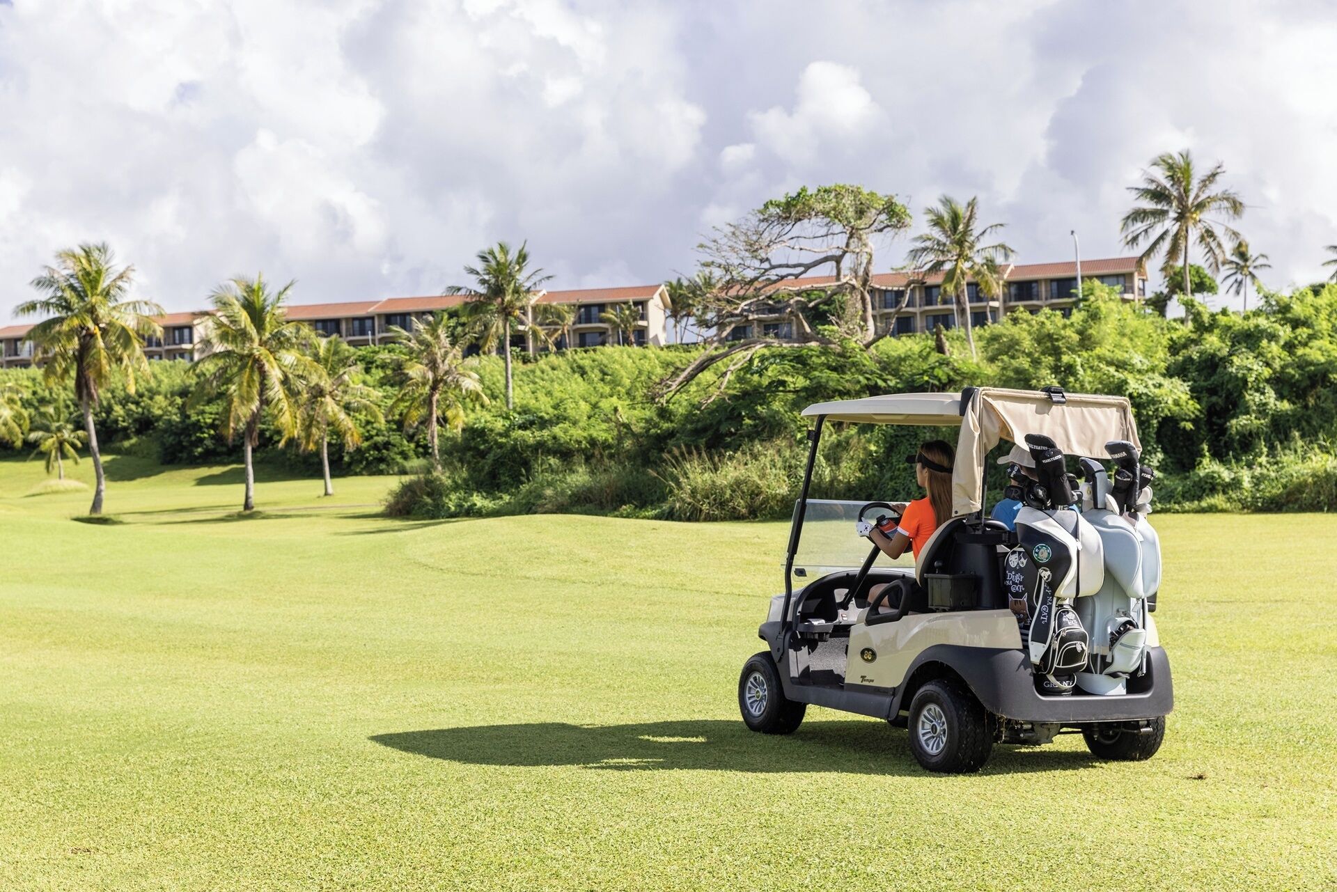 Une voiturette de golf sur un terrain verdoyant bien entretenu, avec en arrière-plan des palmiers et un bâtiment doté d’un toit rouge.