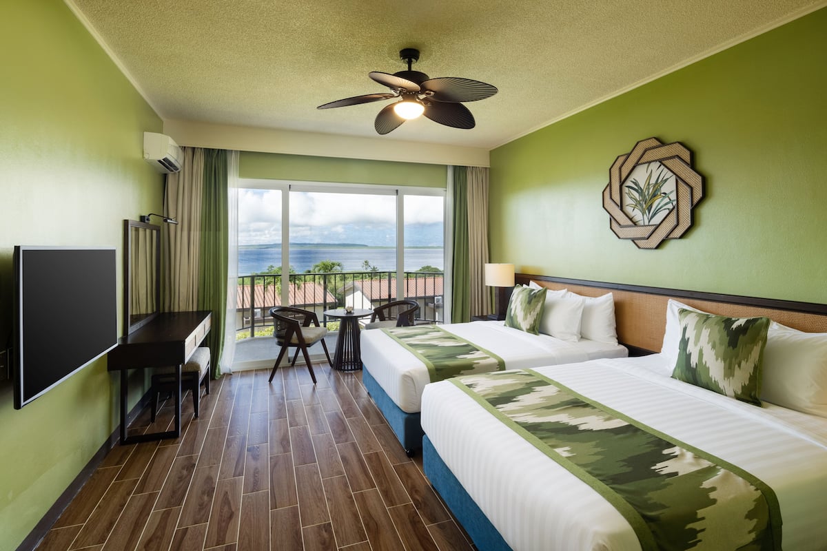 Habitación de hotel con dos camas, televisión, ventilador de techo, vista al océano y balcón con sillas.