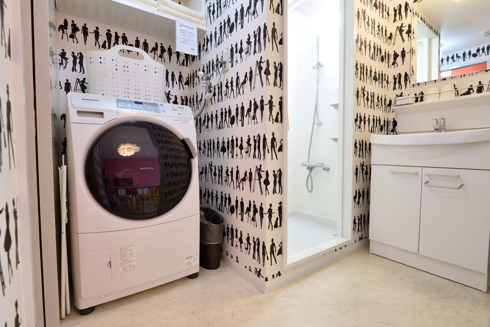 A compact laundry room with a front-loading washing machine, a small trash bin, a shower, and a white cabinet.