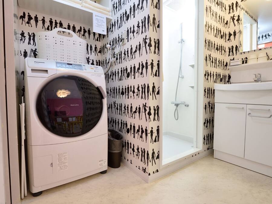 A compact laundry room with a front-loading washing machine, a small trash bin, a shower, and a white cabinet.