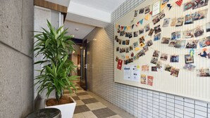 Um corredor com chão xadrez, um vaso de planta, lixeiras e um mural com fotos e avisos.