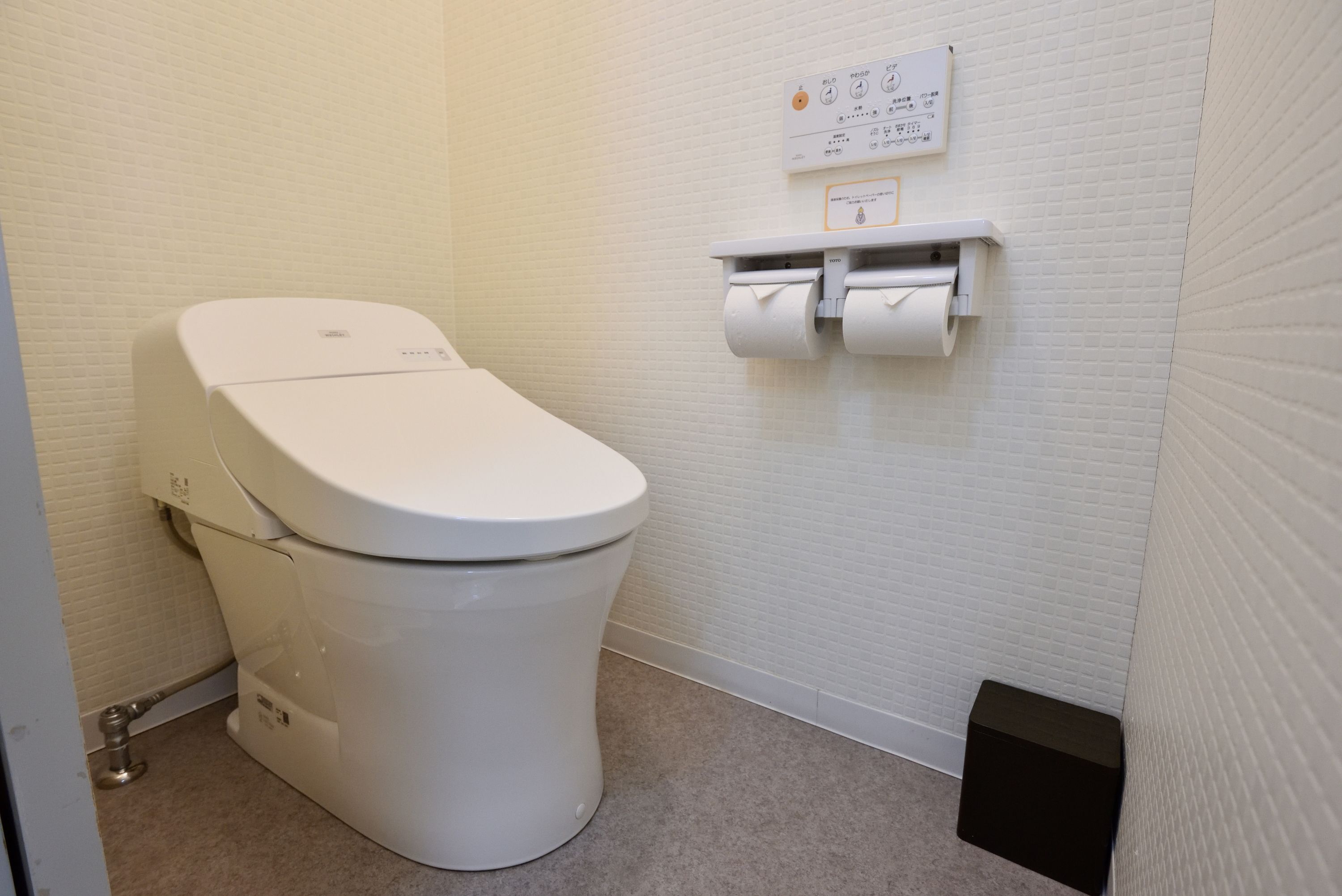 Des toilettes blanches dotées d’un panneau de commande, d’un porte-rouleau de papier toilette et d’une petite poubelle, dans une salle de bain aux murs blancs à texture.