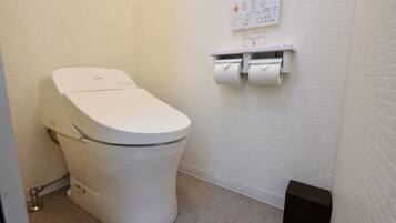 Des toilettes blanches dotées d’un panneau de commande, d’un porte-rouleau de papier toilette et d’une petite poubelle, dans une salle de bain aux murs blancs à texture.