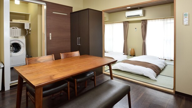 Quarto compacto com mesa de jantar de madeira, um banco, cama com colchão e almofadas, máquina de lavar e guarda-roupa.