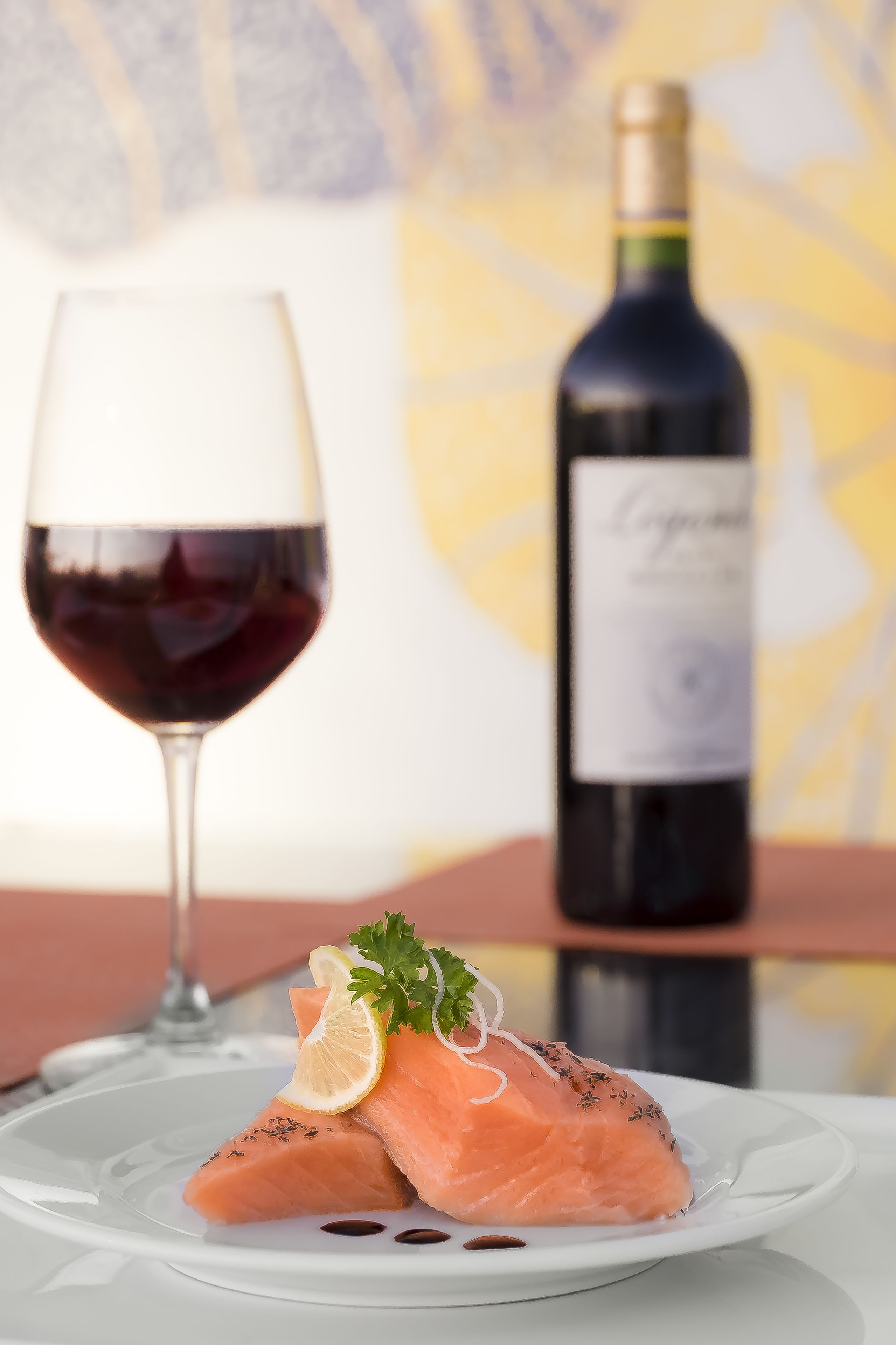 A plate of salmon with lemon and herbs, a glass of red wine, and a bottle of wine on a table.
