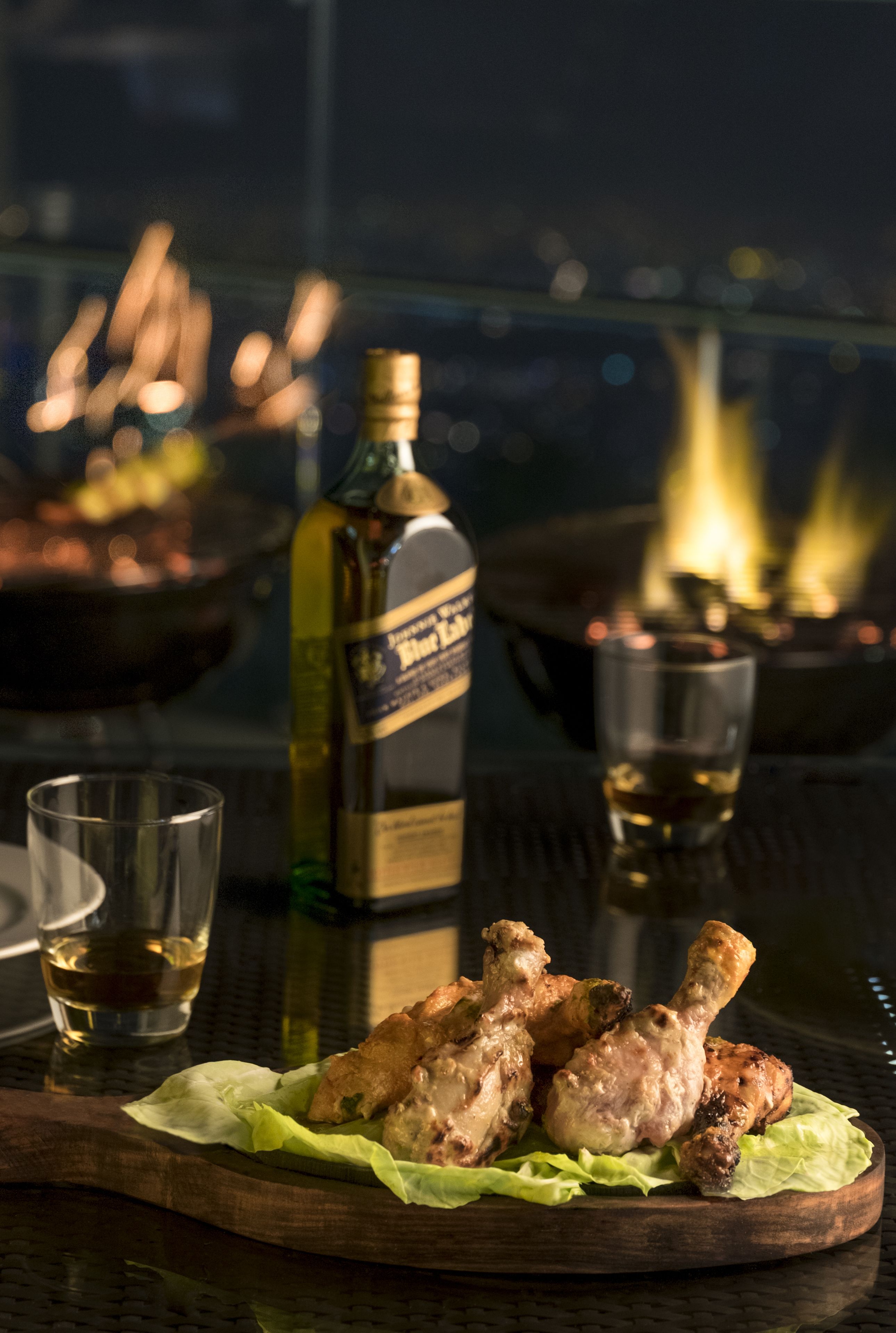 Una tabla de madera con alitas de pollo a la parrilla sobre lechuga, una botella de whisky y un vaso de whisky sobre una mesa con superficie reflectante.