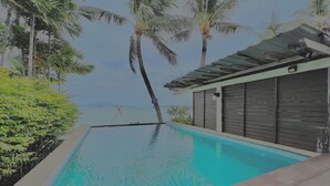 A swimming pool with a view of the ocean, surrounded by palm trees and a building with a sloped roof. - The Shore Samui (Koh Samui)