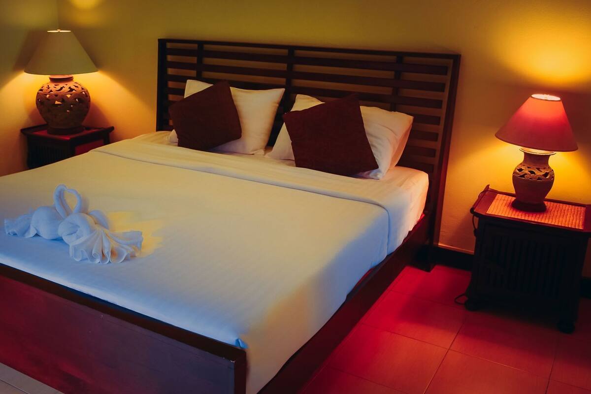 Una cama bien tendida con una toalla y un arreglo de toallas en forma de corazón, dos lámparas de noche y un cabecero de madera.