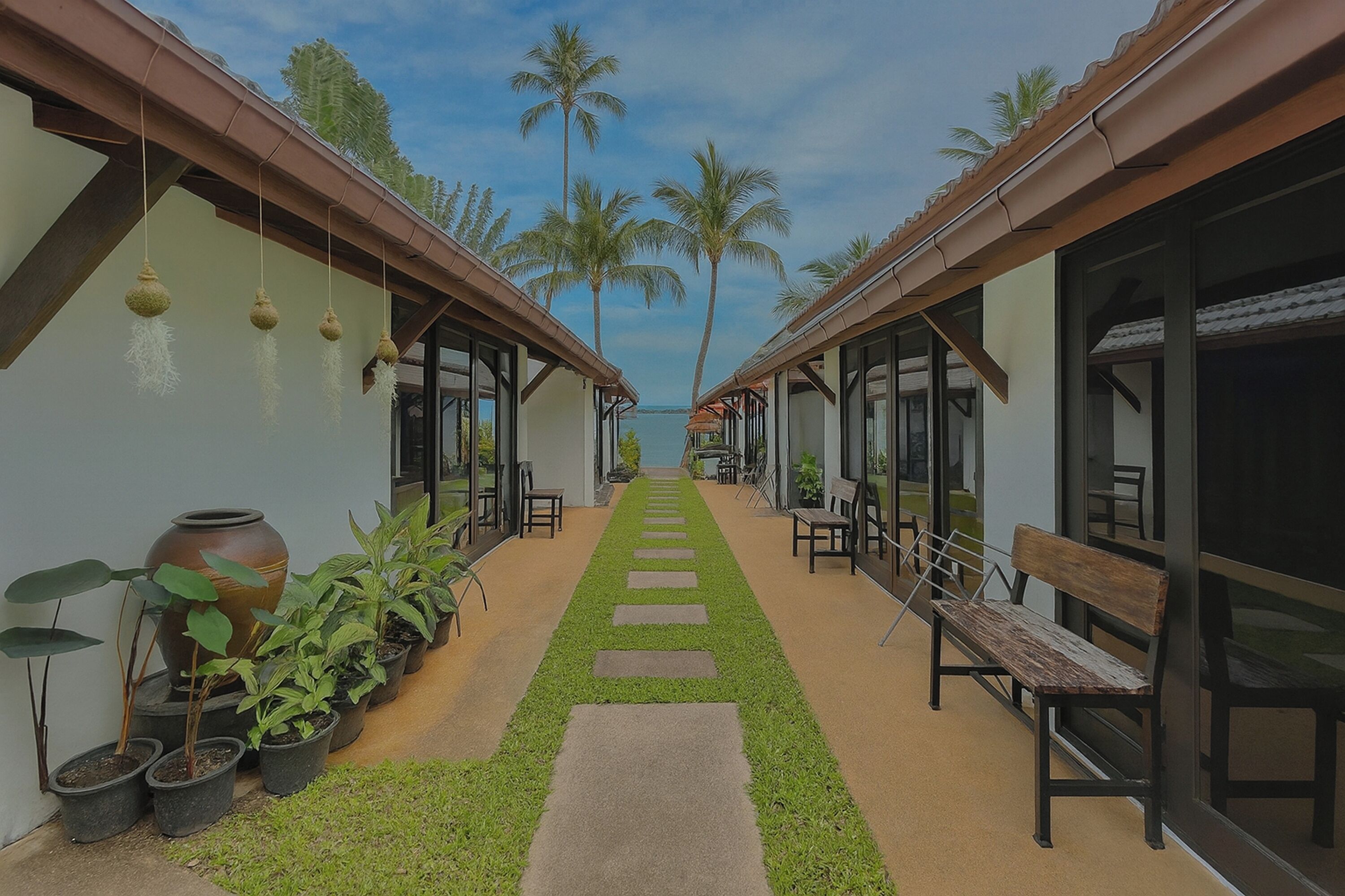 A corridor with a central pathway, benches, potted plants, and a view of palm trees and the sea.