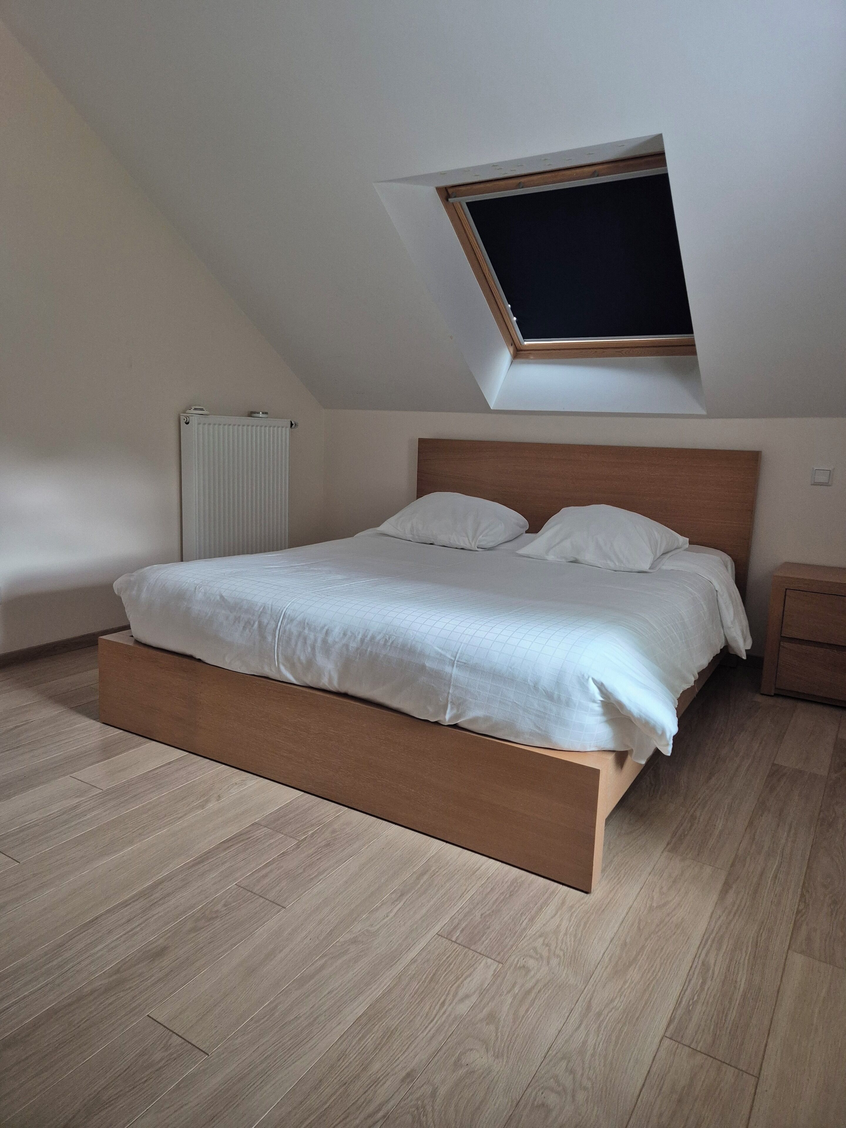 Un dormitorio con techo inclinado, una cama con ropa de cama blanca, cabecero de madera, mesita de noche y un radiador.