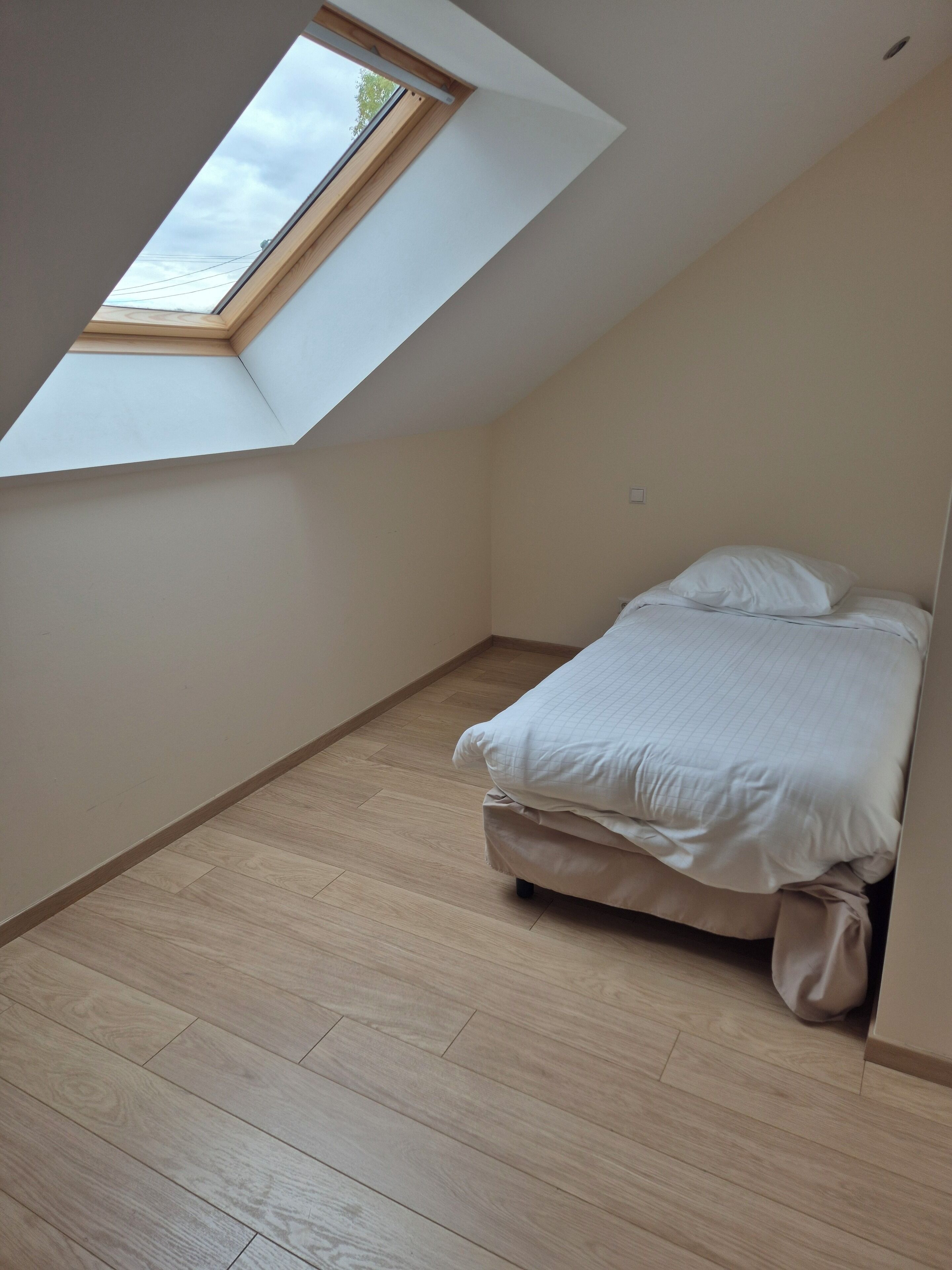 Una habitación pequeña con techo inclinado, tragaluz, cama y suelo de madera.