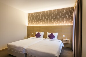 A double bed with white linens and purple pillows, a wooden headboard, a nightstand with a lamp, and a wall-mounted light fixture.