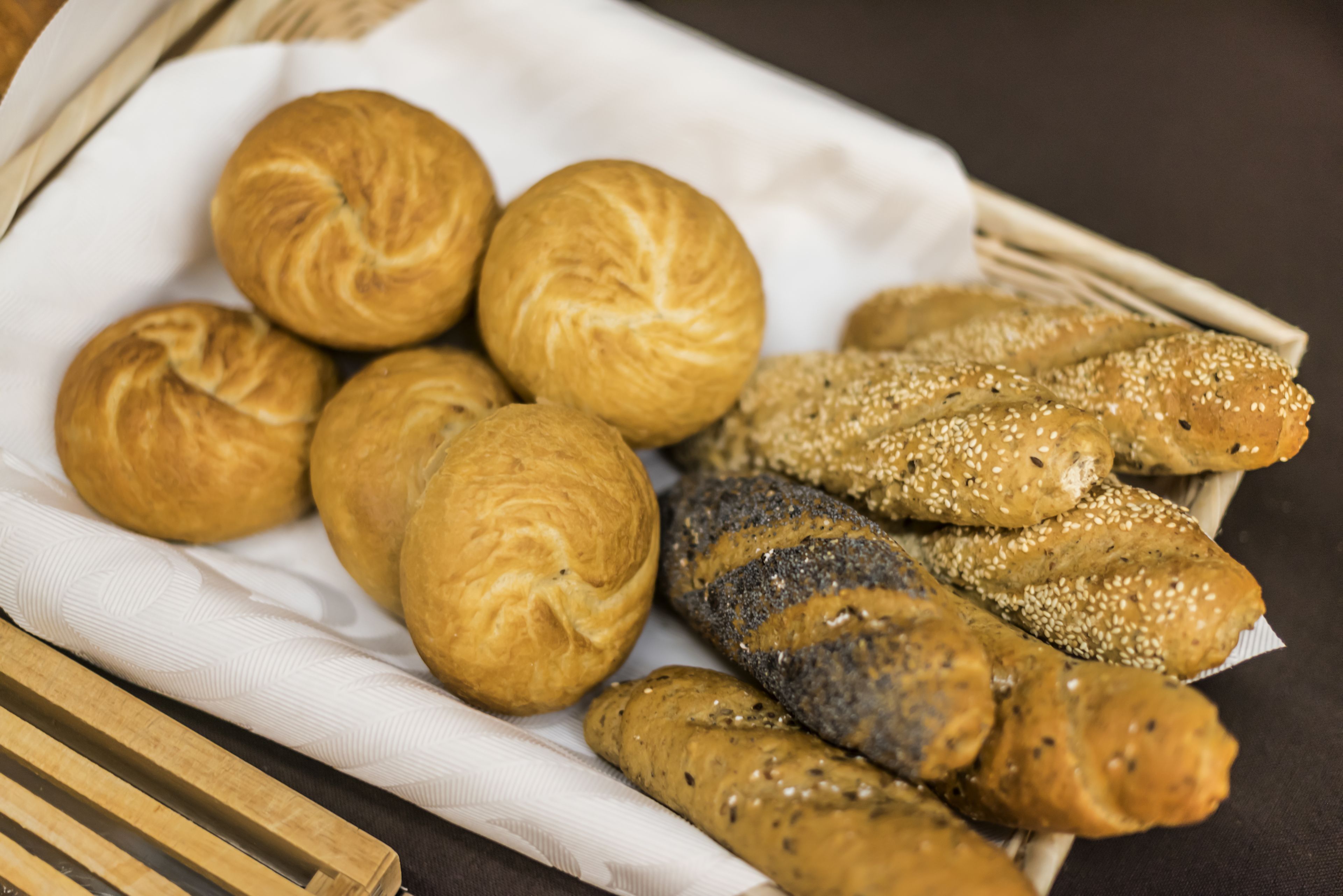 A basket of various breads, including rolls and sesame seed loaves, on a white cloth.