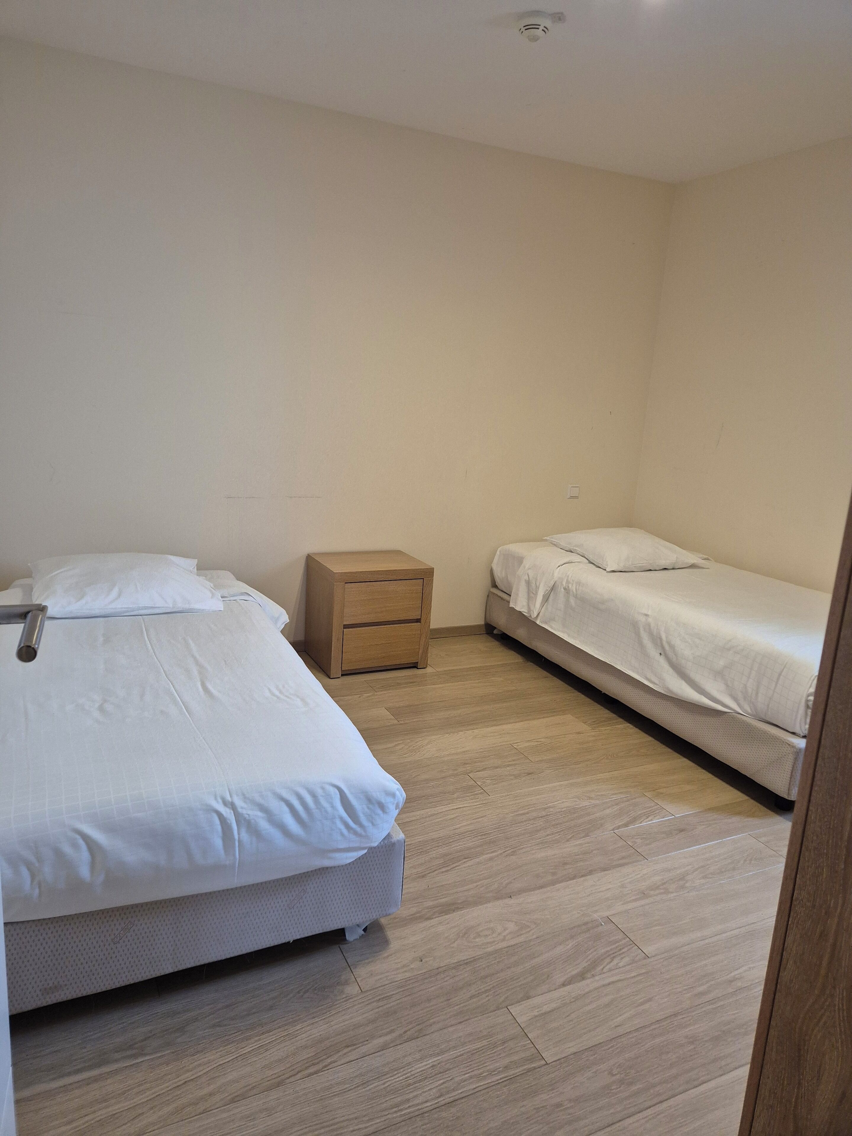 A small, clean room with two single beds, a wooden nightstand, and a light-colored wooden floor.