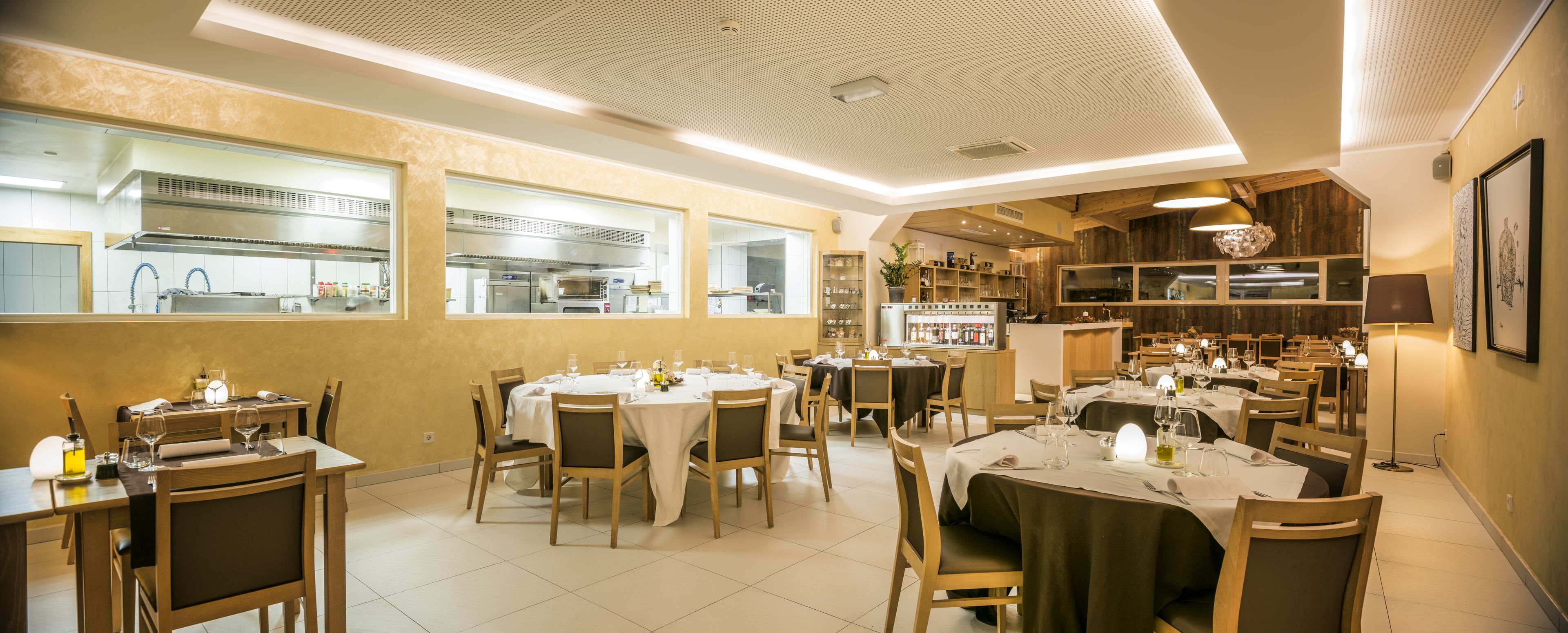 En restaurant med flere spiseborde, stole og et køkkenområde, der kan ses gennem store vinduer.