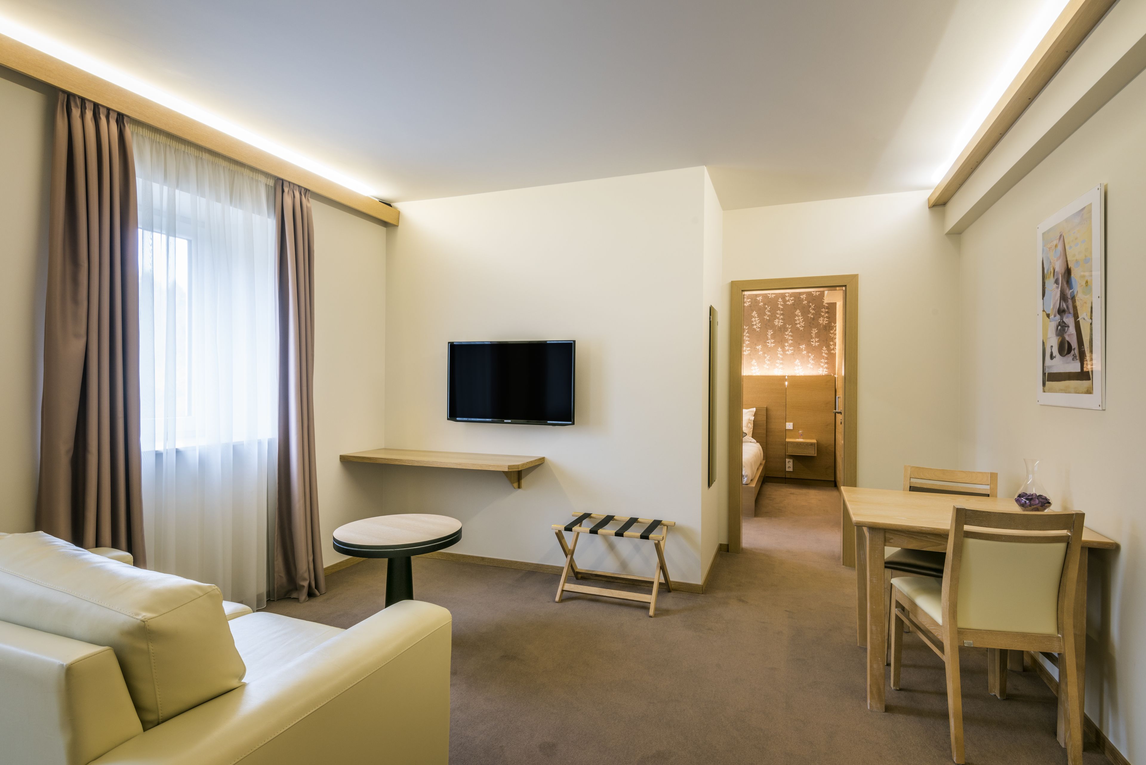 Quarto de hotel com sofá, mesa redonda pequena, TV fixada na parede, um banco de madeira e um espaço de refeições com mesa e cadeiras.