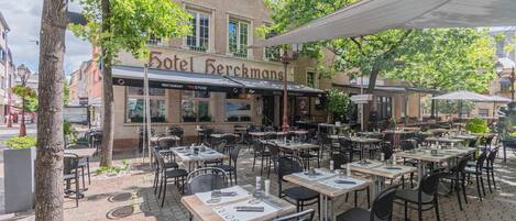 An outdoor seating area with tables and chairs, a building with a sign reading 'Hotel Herckmans,' and trees providing shade.