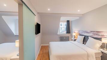 A hotel room with a bed, a desk, a TV, a window with a view of buildings, and a radiator.