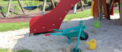 Un parque infantil con un tobogán rojo, una pala azul y una regadera amarilla.