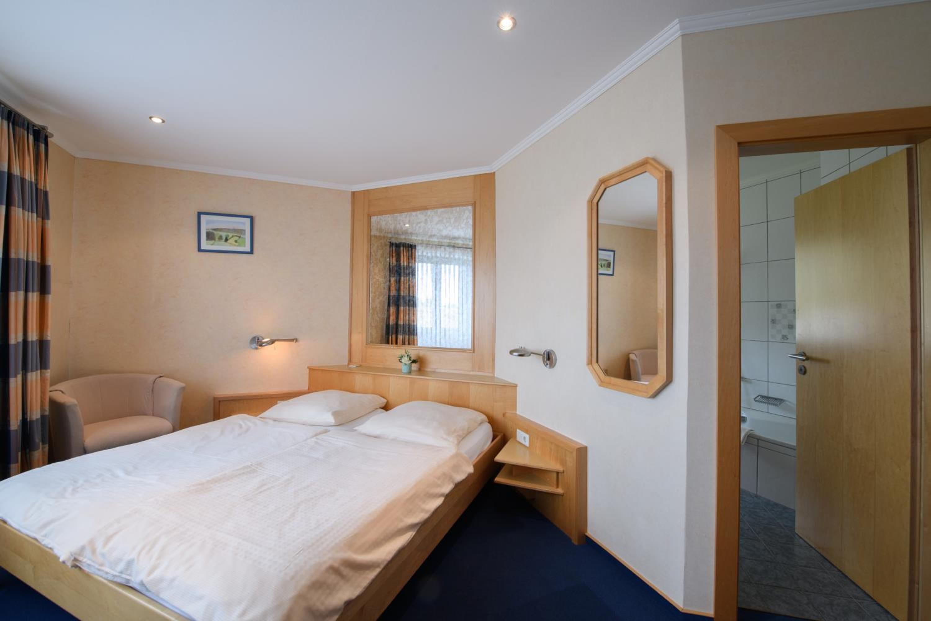 Een hotelkamer met een tweepersoonsbed, een stoel, een nachtkastje, een spiegel en een deur naar een badkamer.