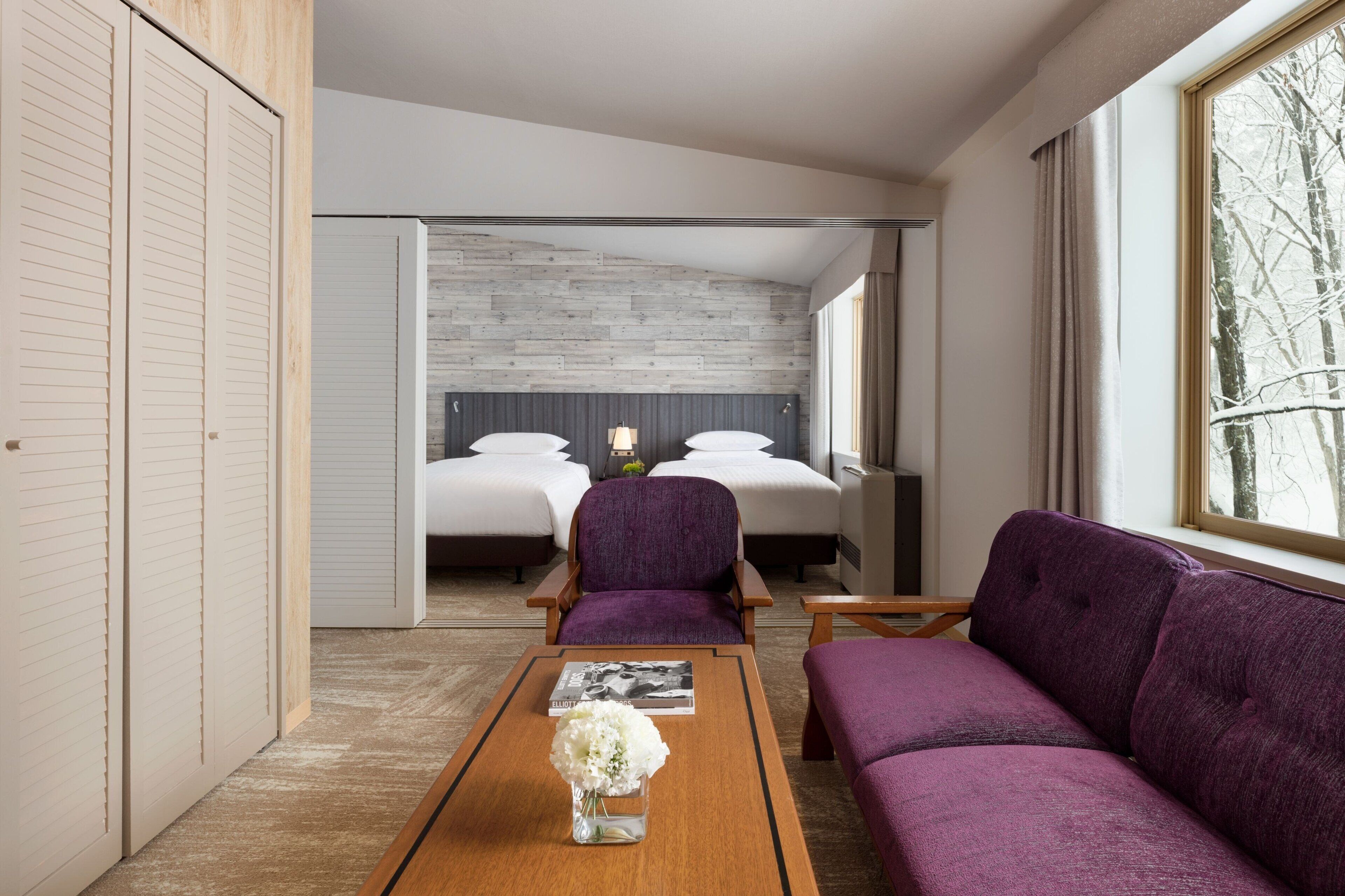 Moderni hotellihuone, jossa on puulattia, violetti sohva, puinen sohvapöytä ja valkoisilla lakanoiden kanssa varustettu sänky.