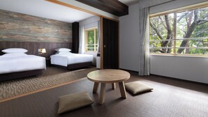 A hotel room with two beds, a round wooden table, and a large window with a view of trees.