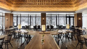 A spacious dining area with wooden tables and chairs, large windows with a snowy view, and a wooden ceiling with hanging light fixtures.