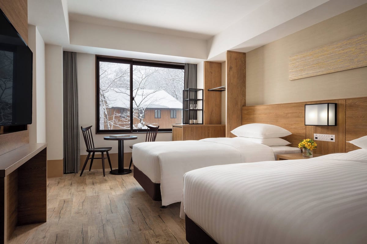 A hotel room with two beds, a desk, a chair, a TV, and a window with a view of a building outside.