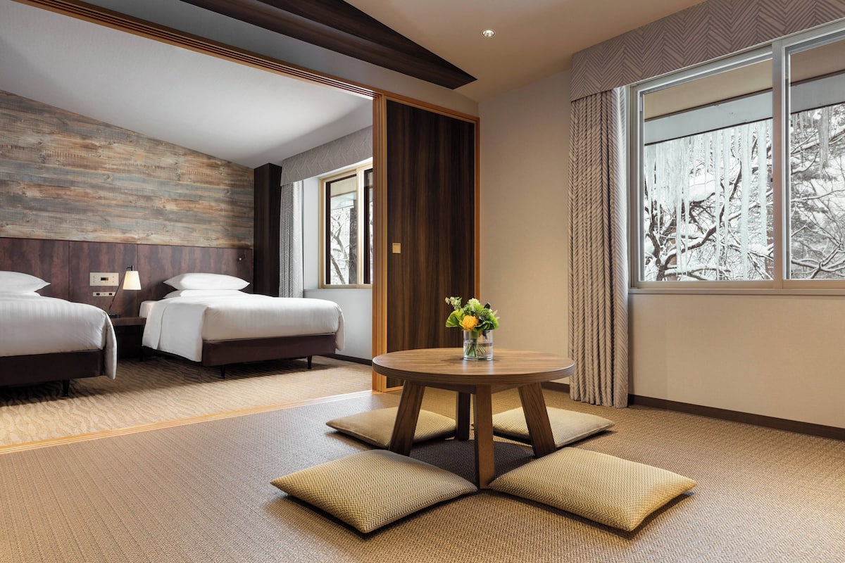 A hotel room with two beds, a round wooden table with a vase of flowers, and a view of trees through the window.