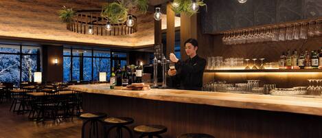 A bar area with wooden bar counter, stools, hanging plants, and a bartender.