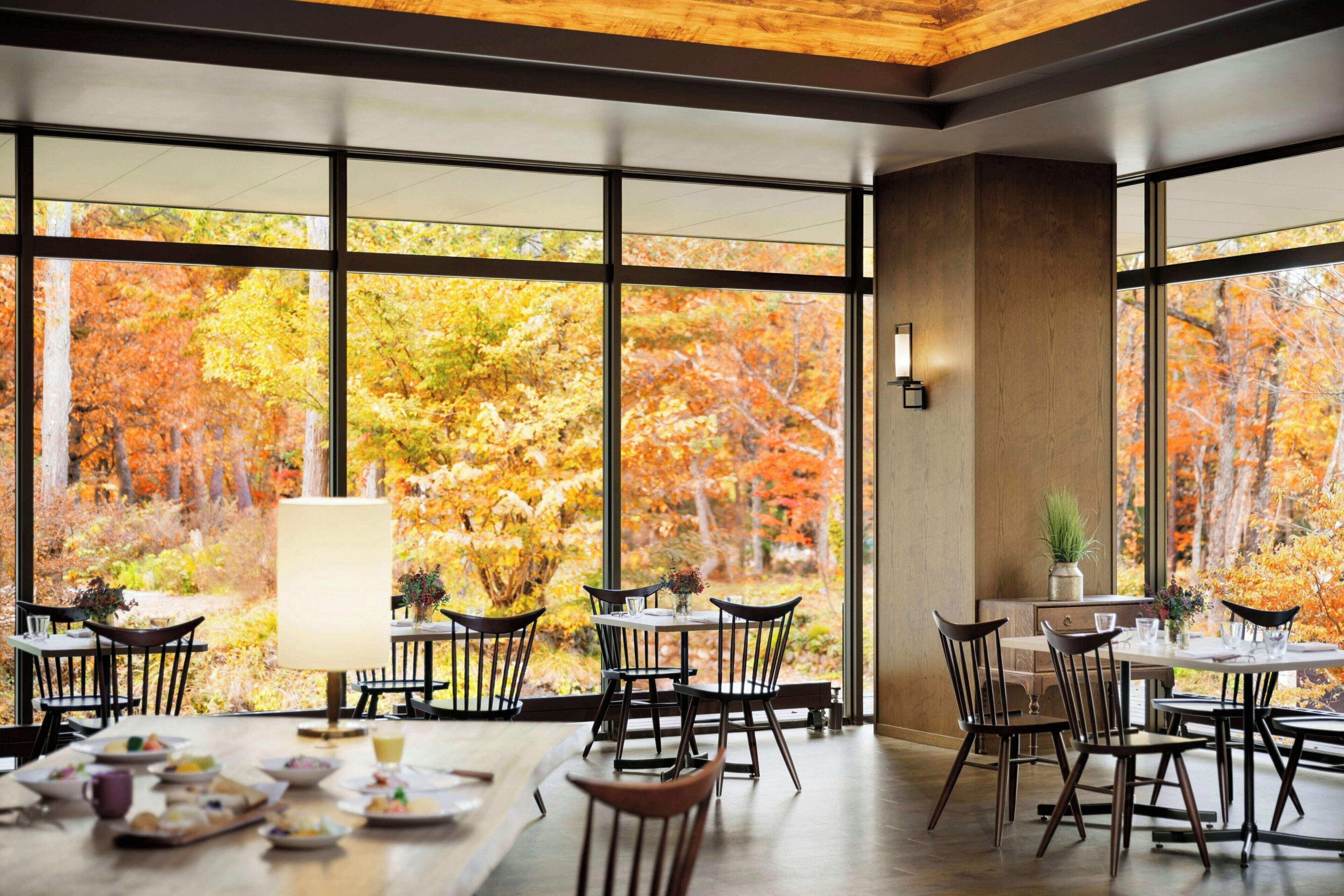 A dining area with large windows offering a view of autumn foliage. Tables are set with plates and glasses.