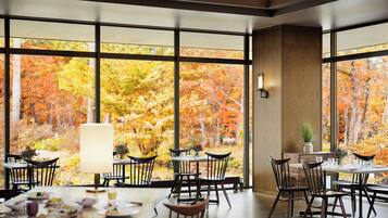 A dining area with large windows offering a view of autumn foliage. Tables are set with plates and glasses.