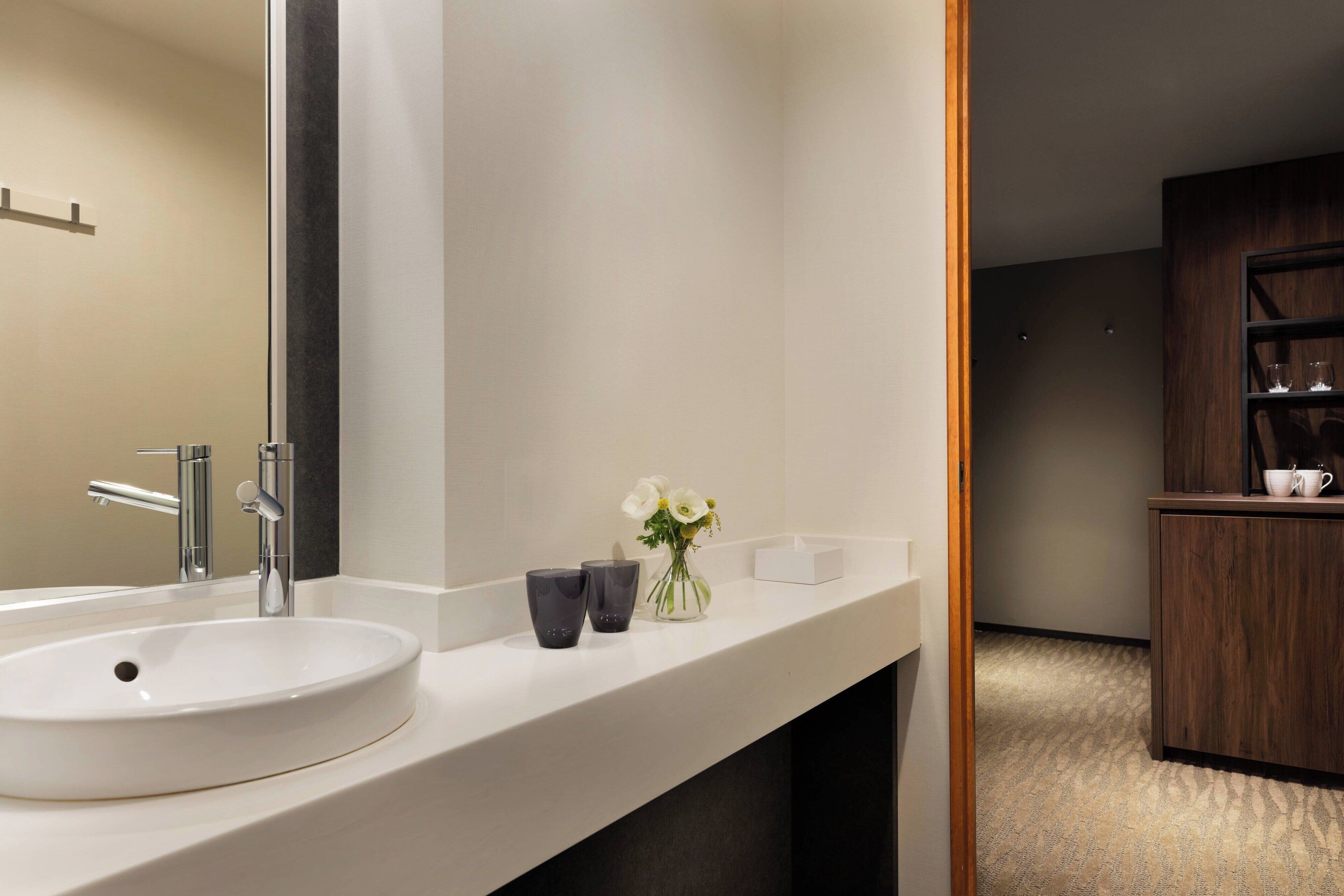 A modern bathroom with a white sink, a large mirror, and a small vase with flowers.
