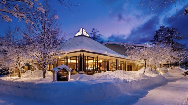 A snow-covered building with a prominent triangular roof and large windows, surrounded by snow-laden trees.