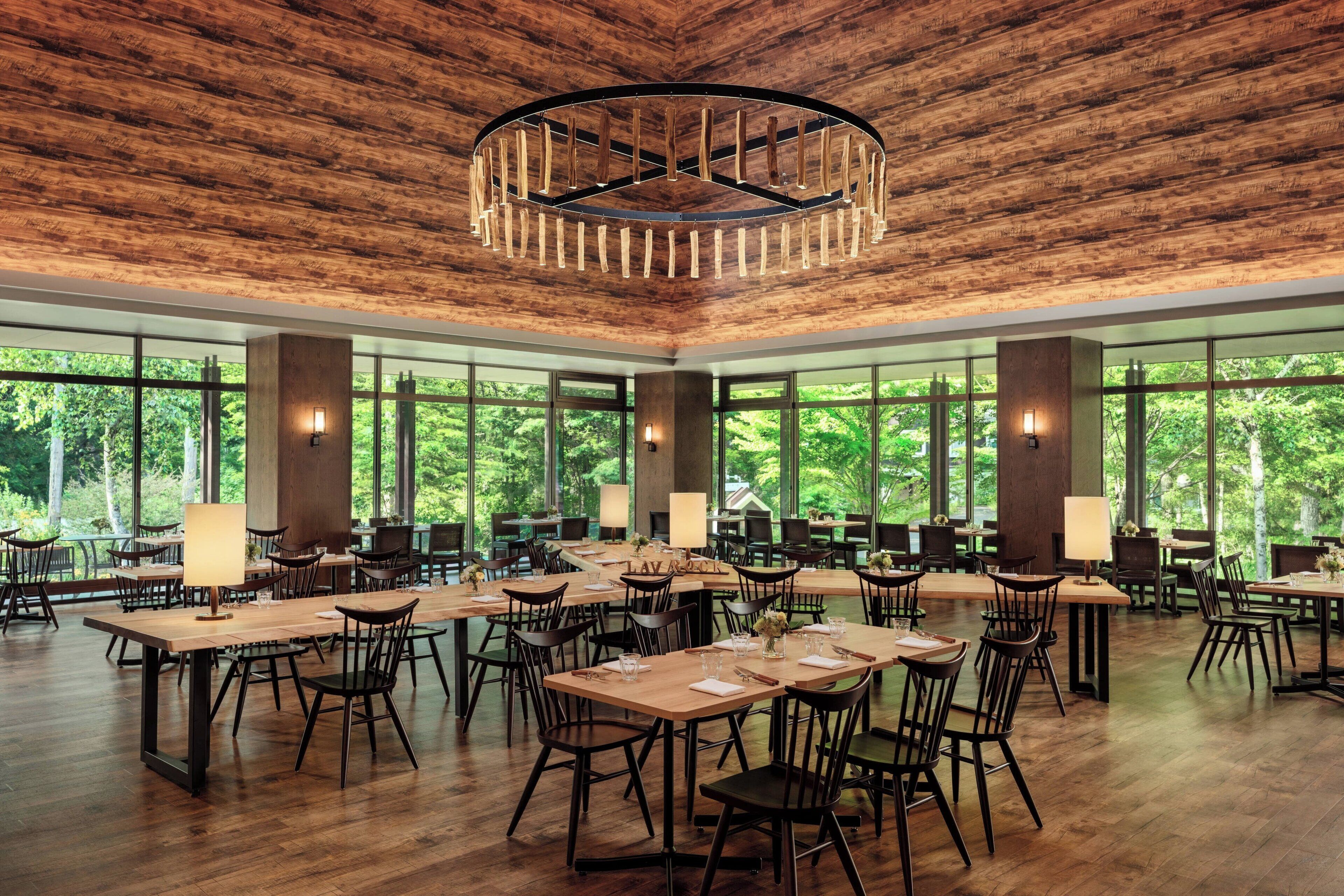A spacious dining area with multiple tables and chairs, large windows offering a view of the outdoors, and a central wooden ceiling with a circular light fixture.