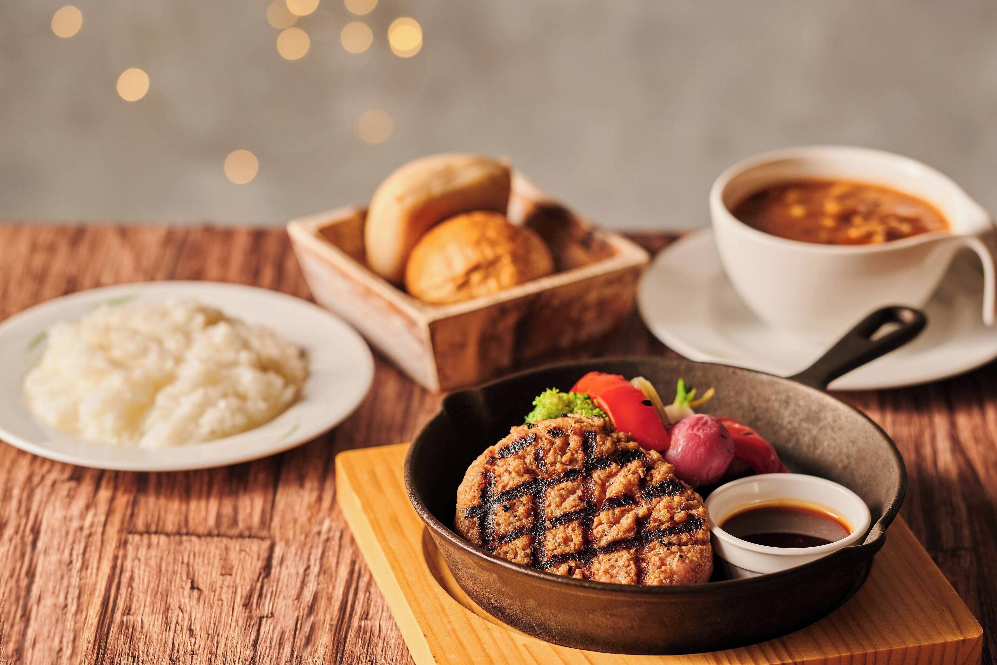 Un plato con carne a la parrilla, puré de papas y una taza de sopa sobre una mesa de madera.