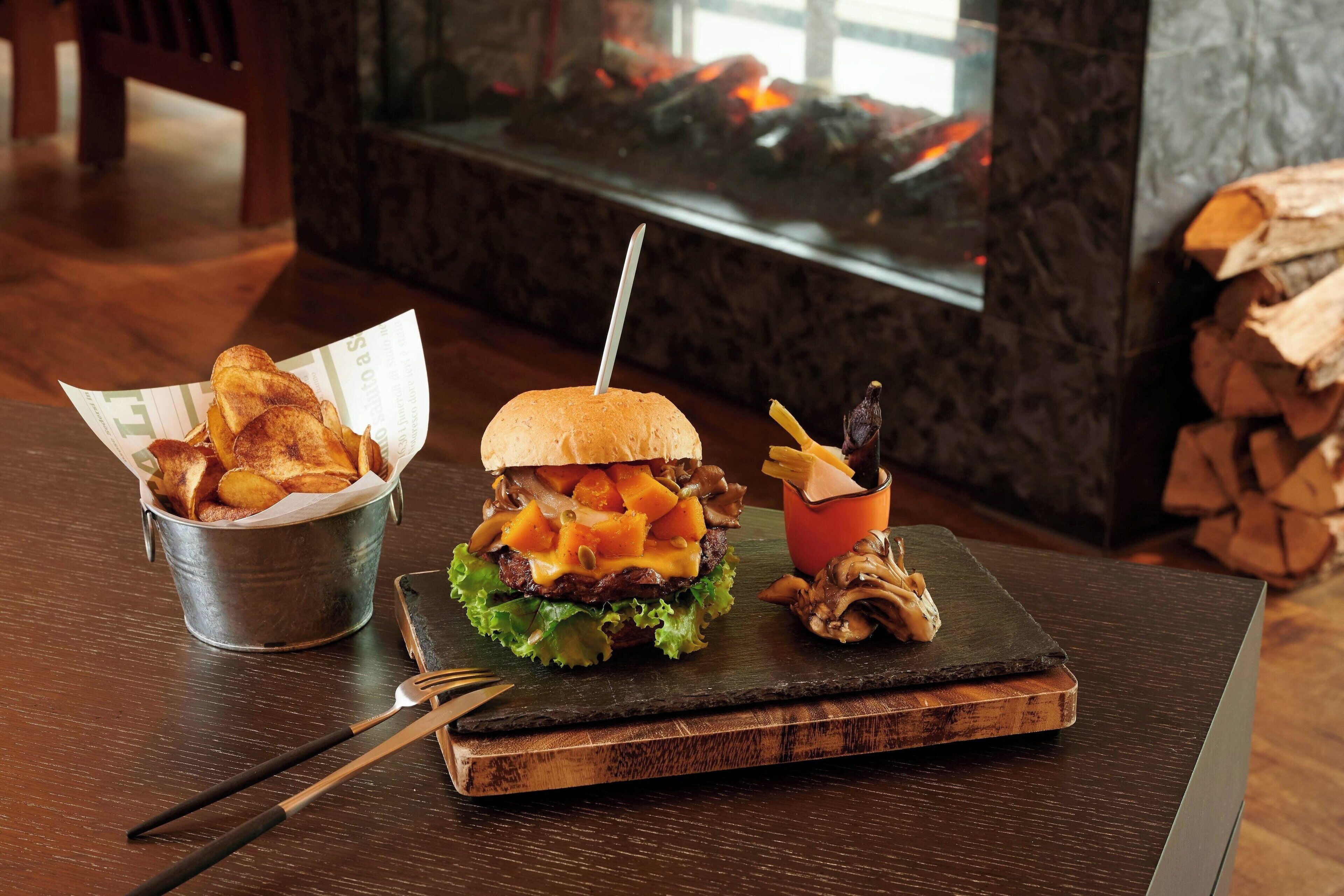 A burger with fries and mushrooms on a wooden board, with a fireplace in the background.