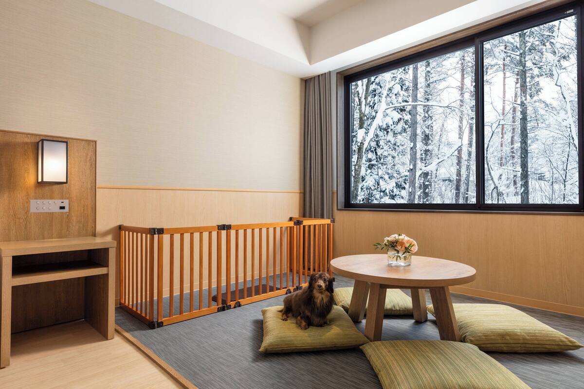 A modern room with a wooden crib, a small table with a vase of flowers, and a large window with a snowy forest view.